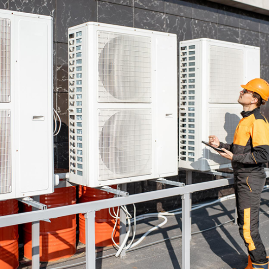 Technician in safety gear inspecting large industrial air conditioning units outside a building.